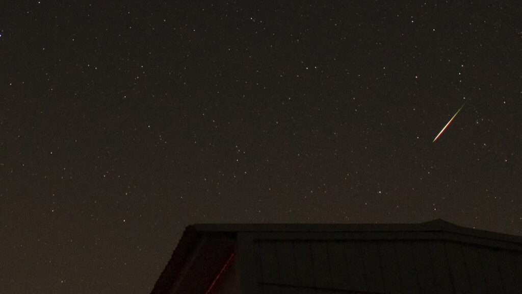 A short, bright, colorful Perseid meteor. The silhouette of the roof of a shed is in the foreground.