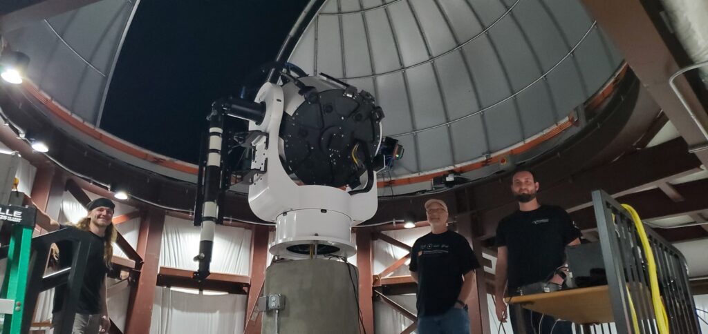 Three men stand near a telescope mounted on top of a tall pier inside an observatory dome.