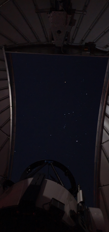 A large telescope points through the open aperture of an observatory dome. The hourglass-shaped constellation of Orion is visible in the sky.