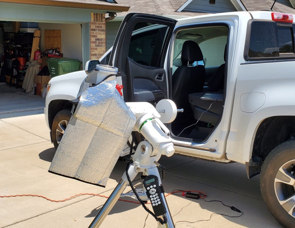 Telescope and mount sitting in place on a paved driveway. A white light solar filter is attached to the objective end of the scope. The scope is covered with a heat shield.